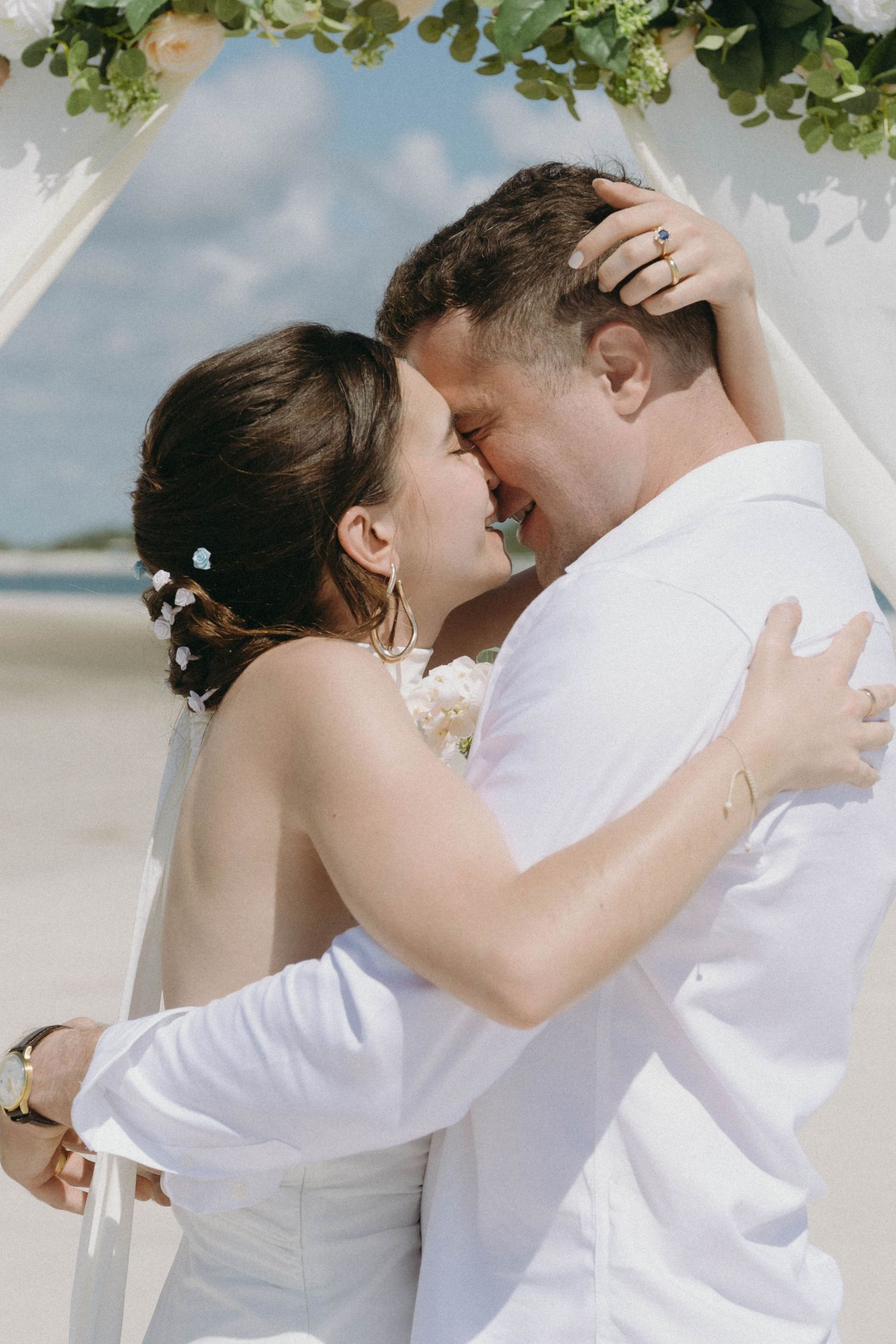 Romantic wedding couple embraces in white attire against a beachy outdoor backdrop.