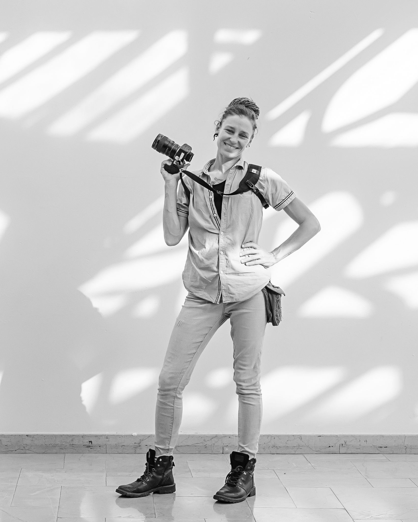 A photographer in casual attire strikes a confident pose with camera equipment against a white wall with dramatic shadows.
