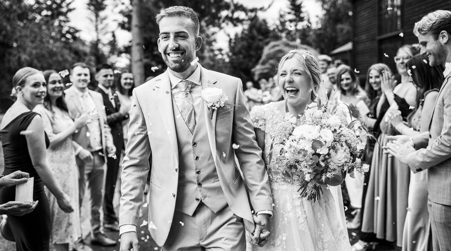Black and white photo of laughing newlyweds walking through confetti shower surrounded by cheering guests.