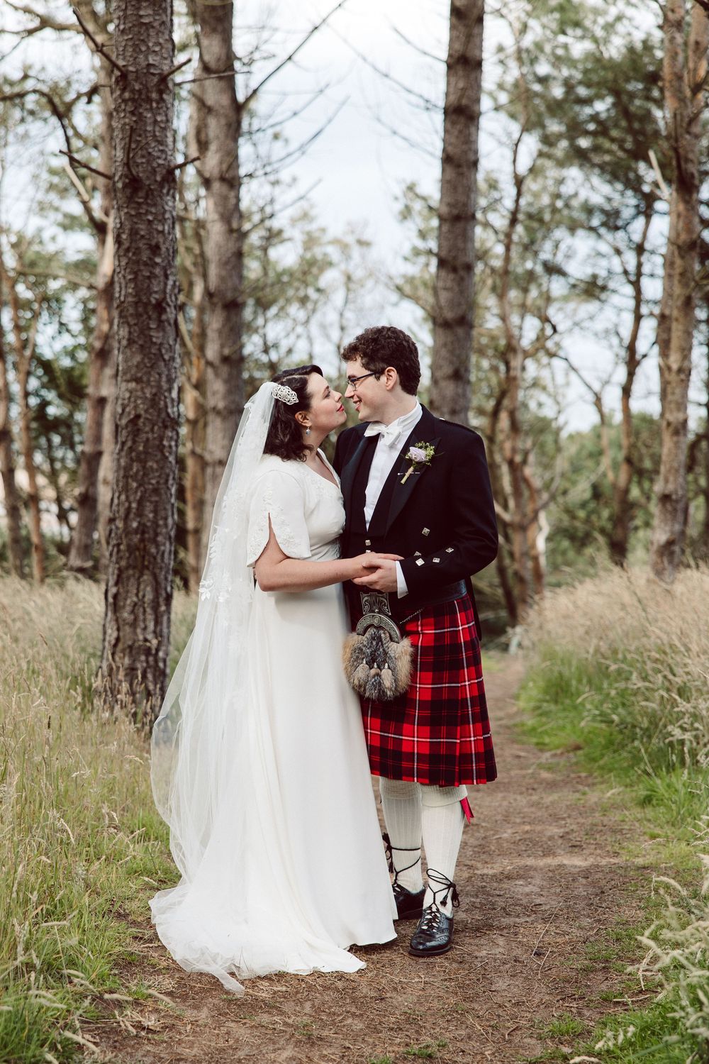 A romantic wedding couple embraces on a wooded path, with the groom wearing a traditional red tartan kilt.