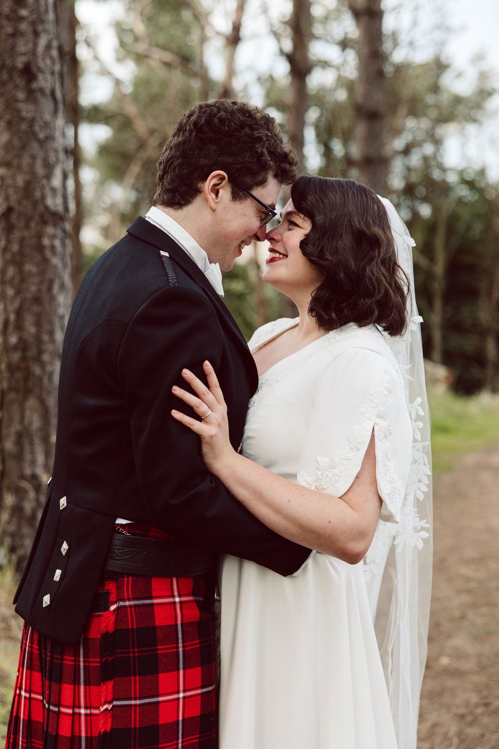 A romantic wedding couple shares a tender moment in a forest setting with the groom wearing a traditional Scottish kilt.