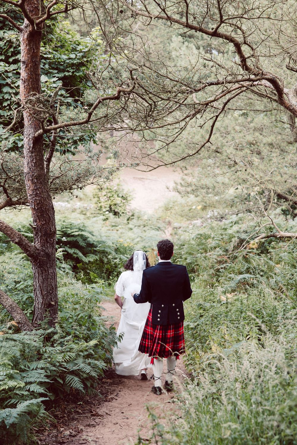 A couple in traditional Scottish wedding attire walking down a wooded forest path together.
