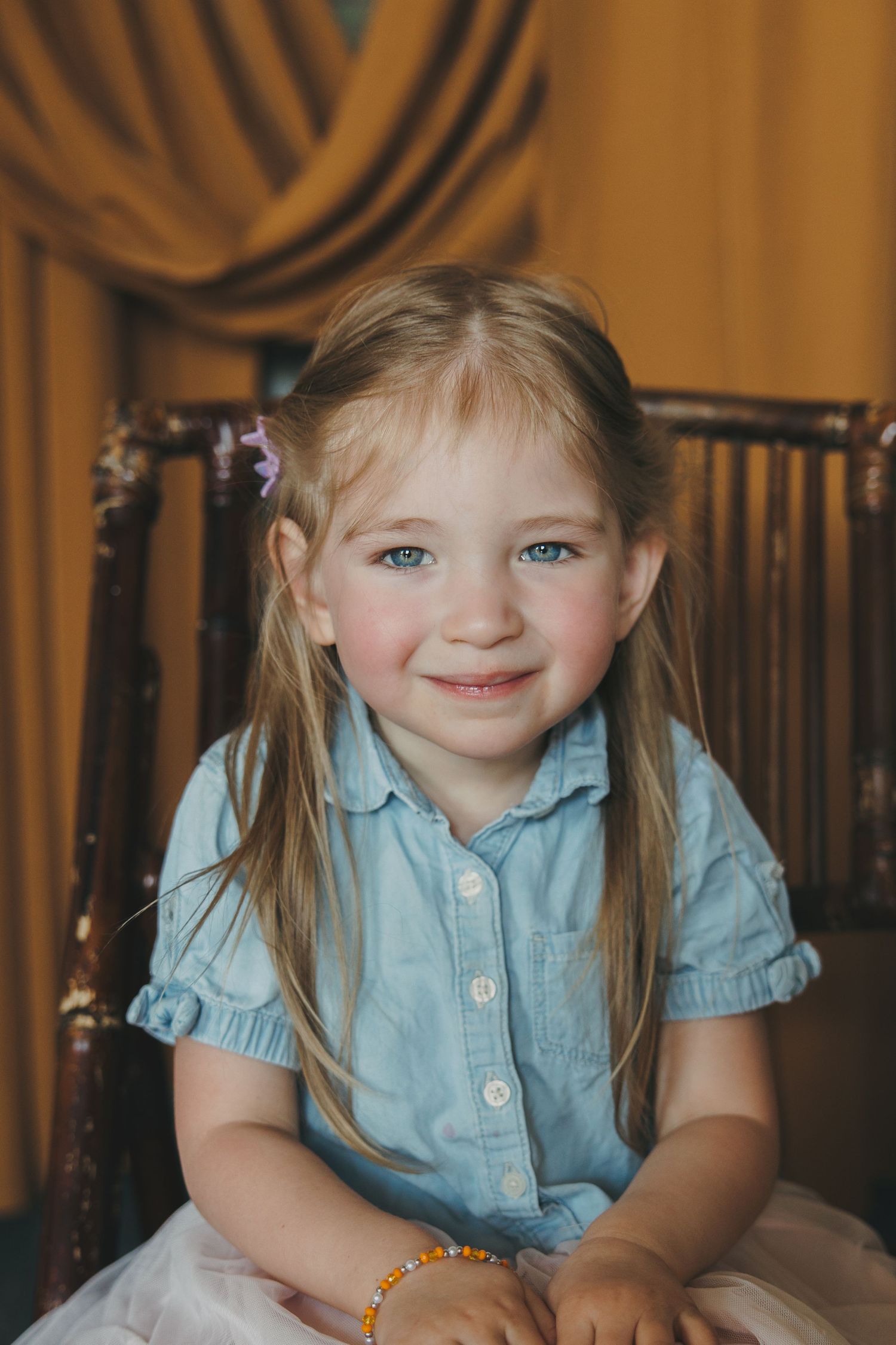 Smiling blonde little girl with blue eyes wearing a blue dress seated in an ornate wooden chair.