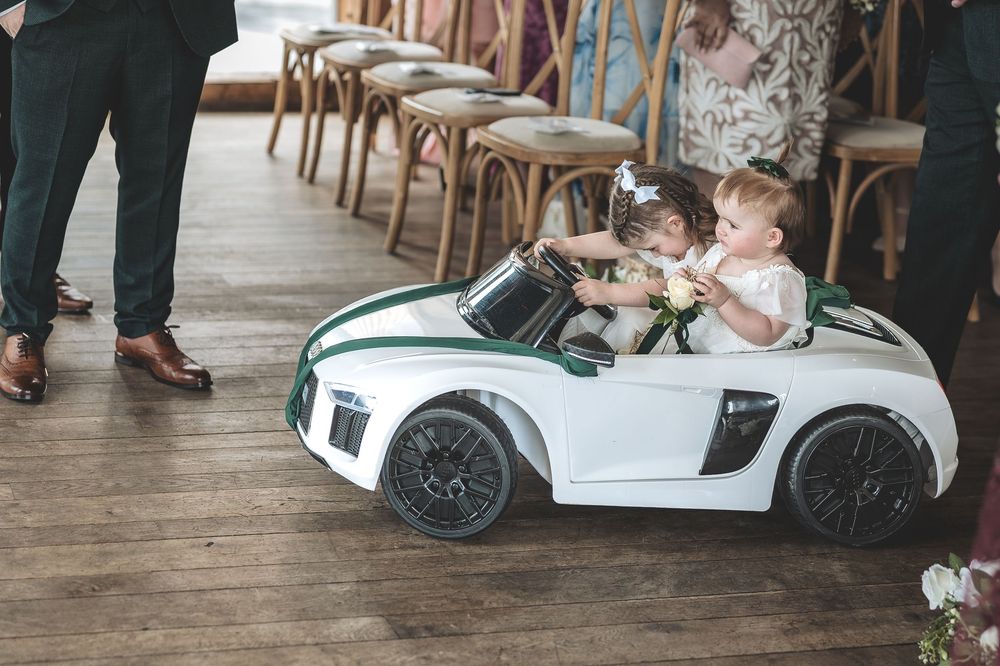 Unobtrusive wedding photography capturing two flower girls in their toy car down the aisle.