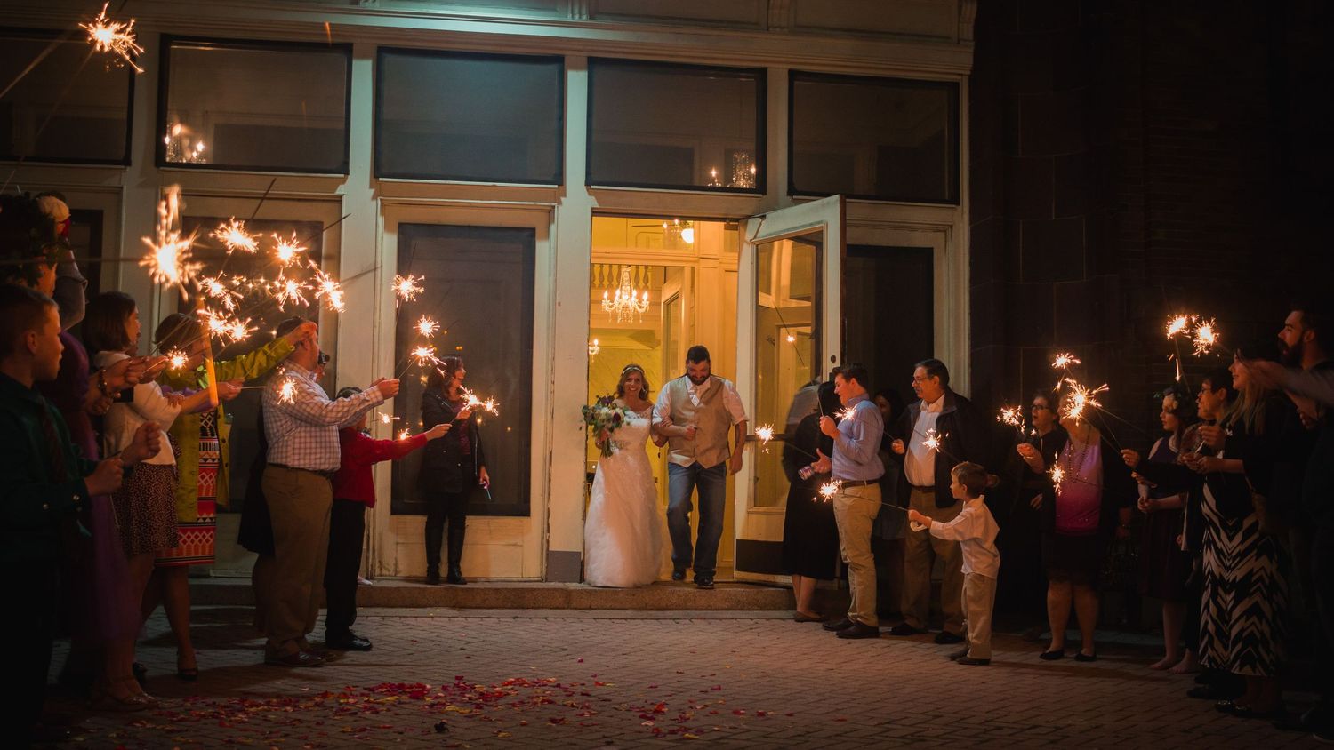 Wedding guests hold sparklers creating a glowing tunnel pathway for the newlyweds during an evening celebration.