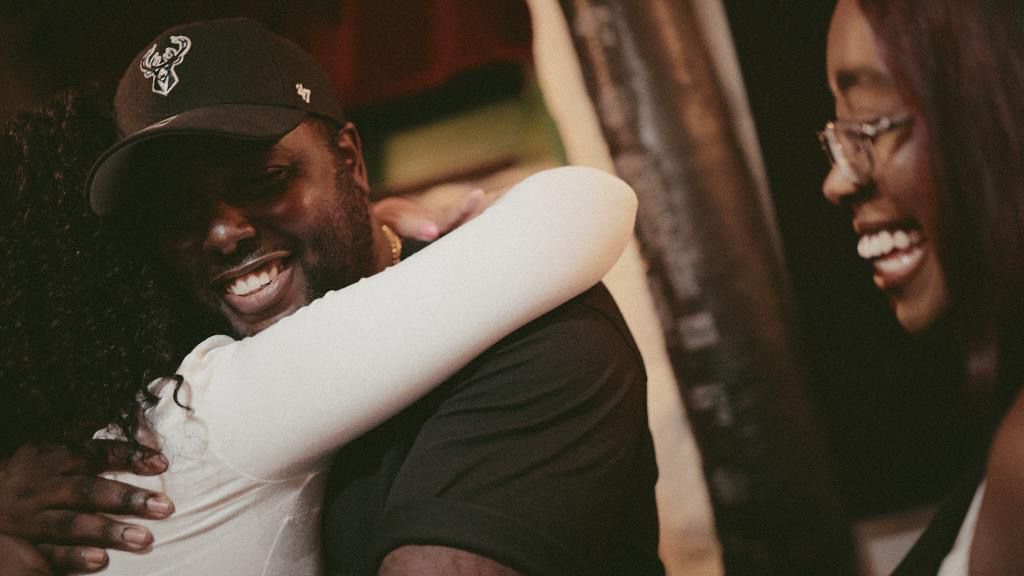 A man in a black cap shares a warm hug with a woman as another woman smiles nearby.