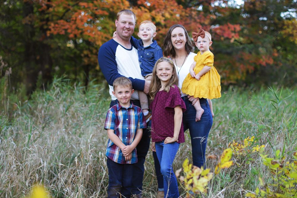 A group poses together outdoors surrounded by vibrant autumn foliage and tall grass in a natural setting.