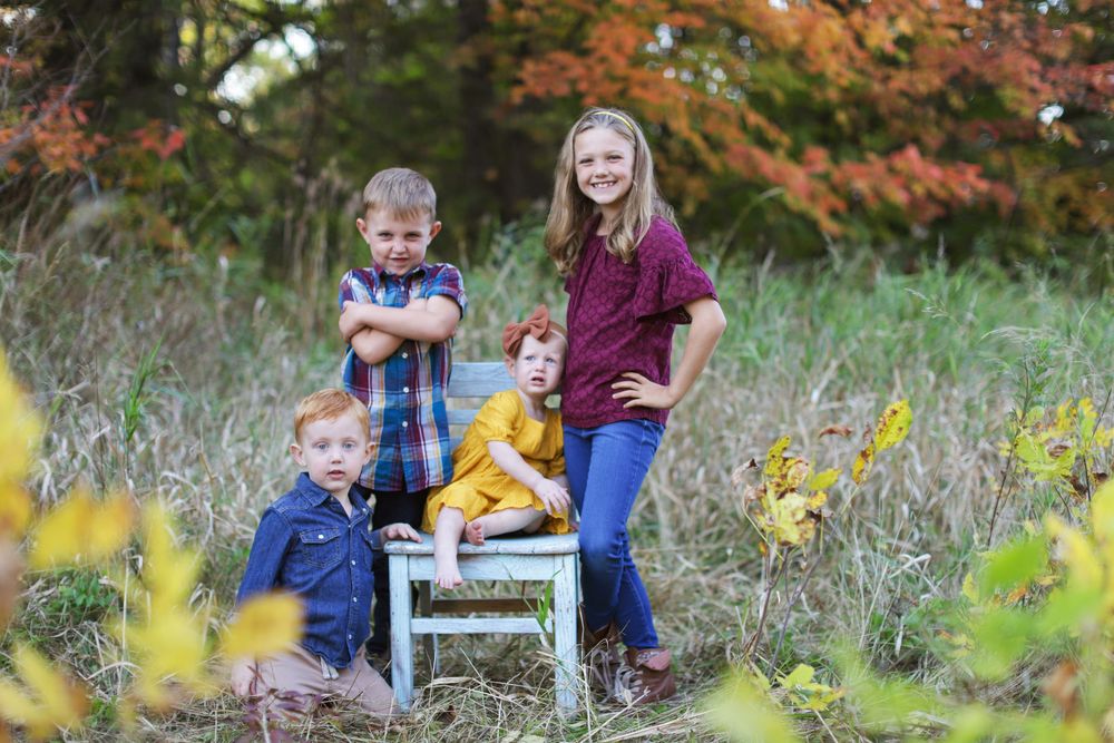 Family enjoys a fall photo session together outdoors among autumn foliage and a vintage white chair.