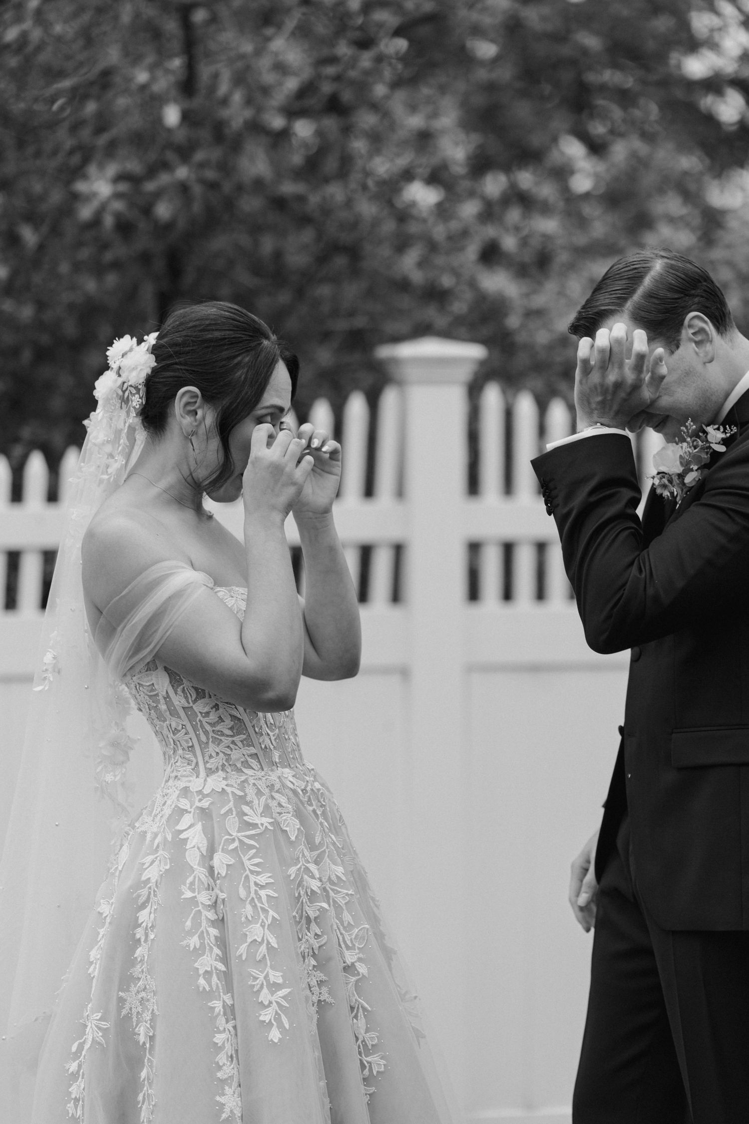 A romantic wedding couple embraces in front of a white picket fence during their outdoor ceremony.
