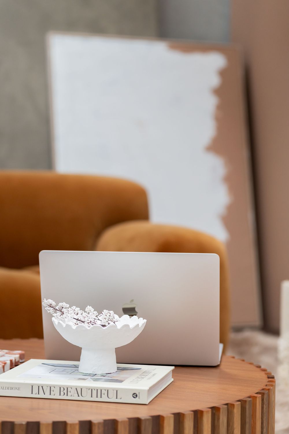 A modern minimalist workspace setup with a wooden desk, MacBook laptop, and white decorative bowl against a neutral background.