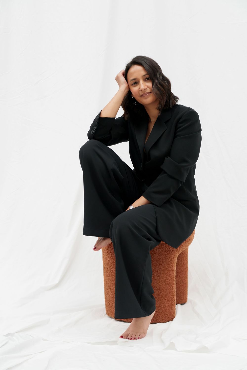 A person wearing a black blazer and pants sits casually on a brown ottoman against a white backdrop in a studio setting.