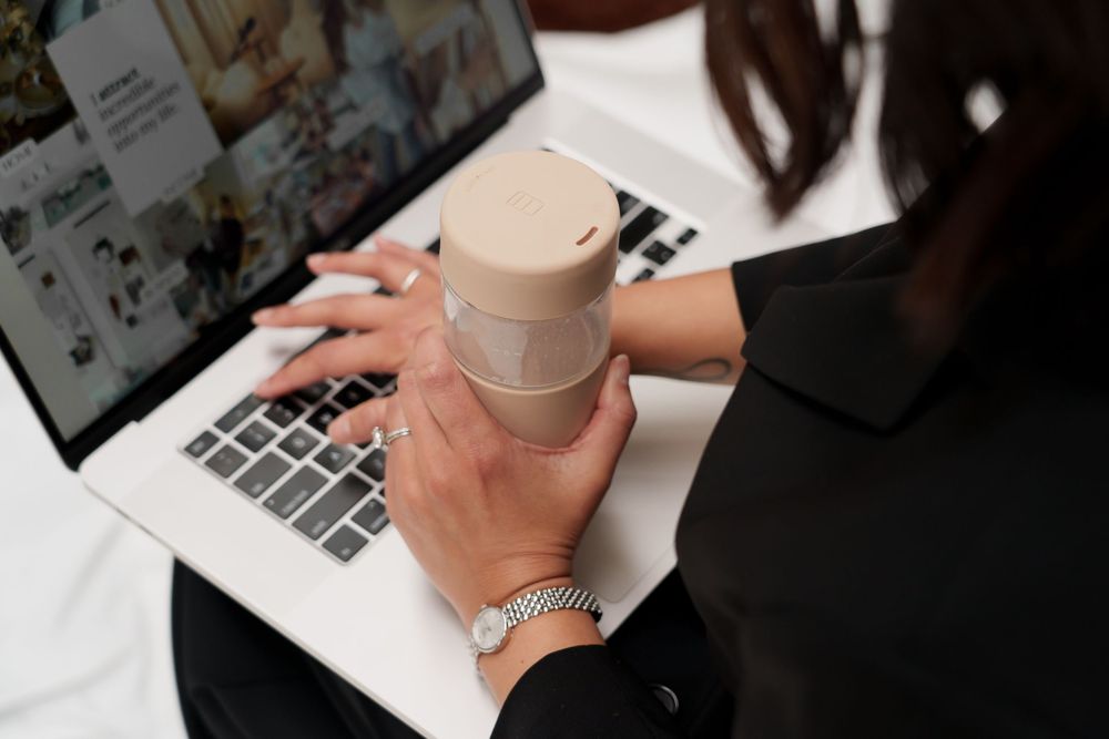 Close-up view of hands typing on laptop keyboard while holding coffee cup in modern workspace setting.
