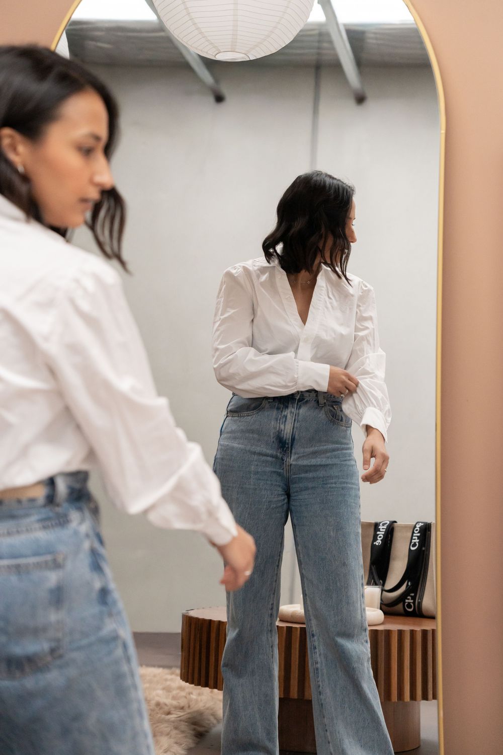 Woman in white button-down shirt and blue jeans poses casually in front of white wall with paper lantern decor.