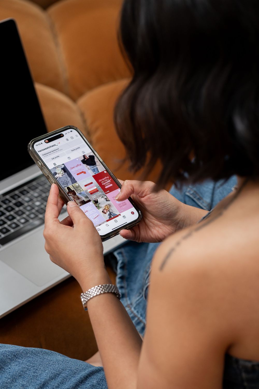 Person scrolling through social media on smartphone while sitting with laptop on brown couch.