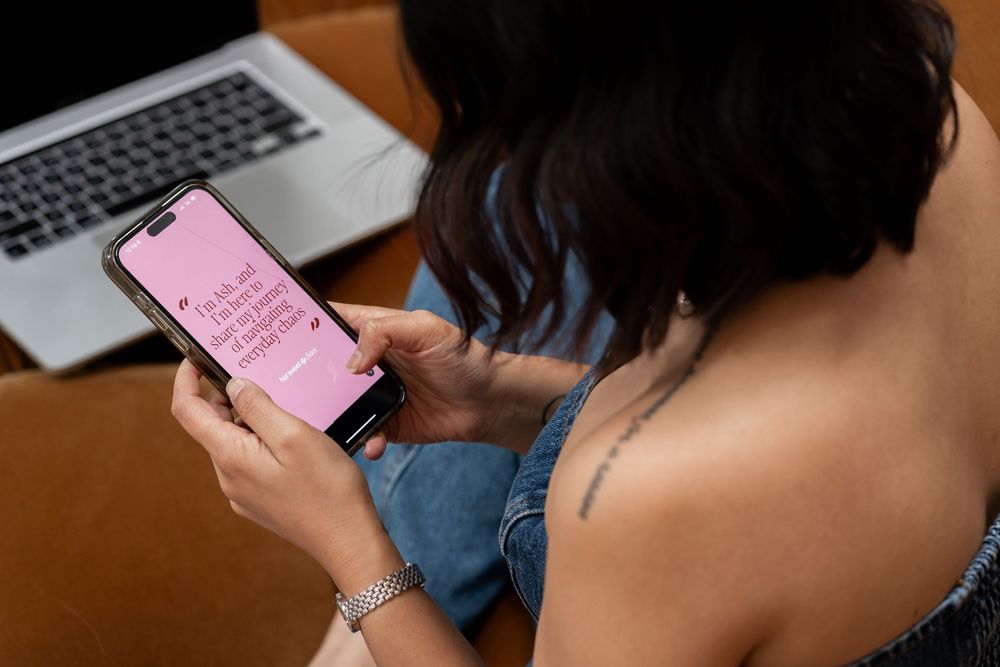 Person using smartphone with pink screen display while sitting at desk with laptop in background.