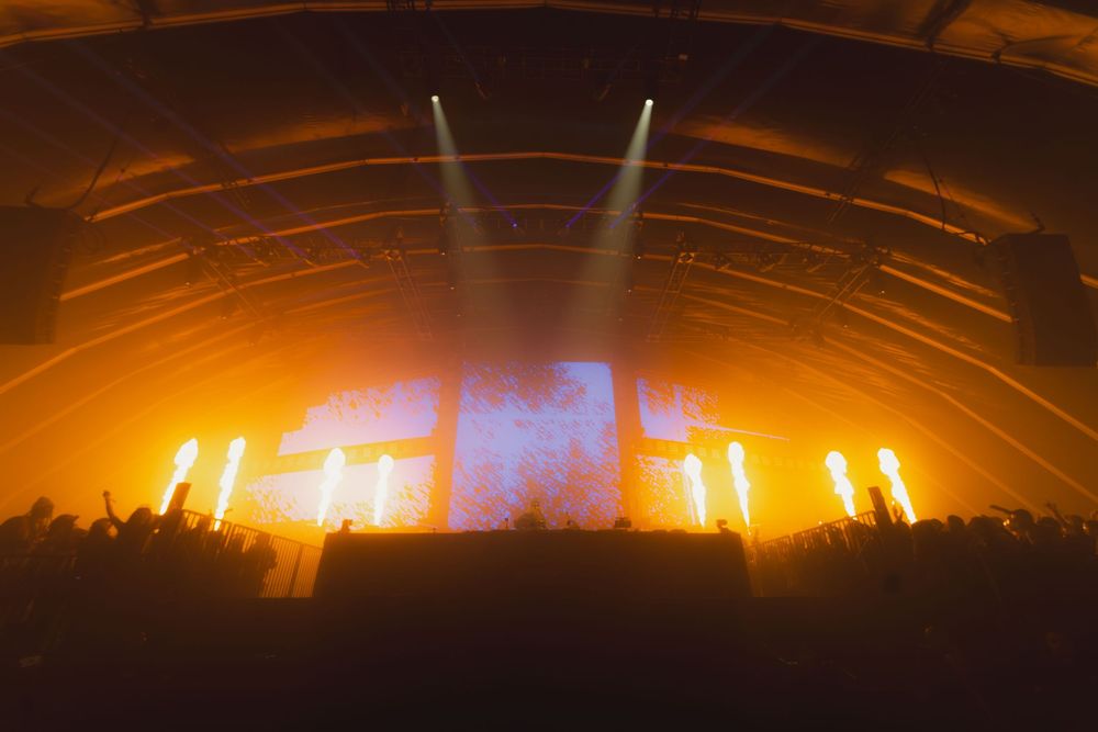 Concert stage illuminated with dramatic orange lighting and blue screen displays with light beams cutting through smoke in darkness.