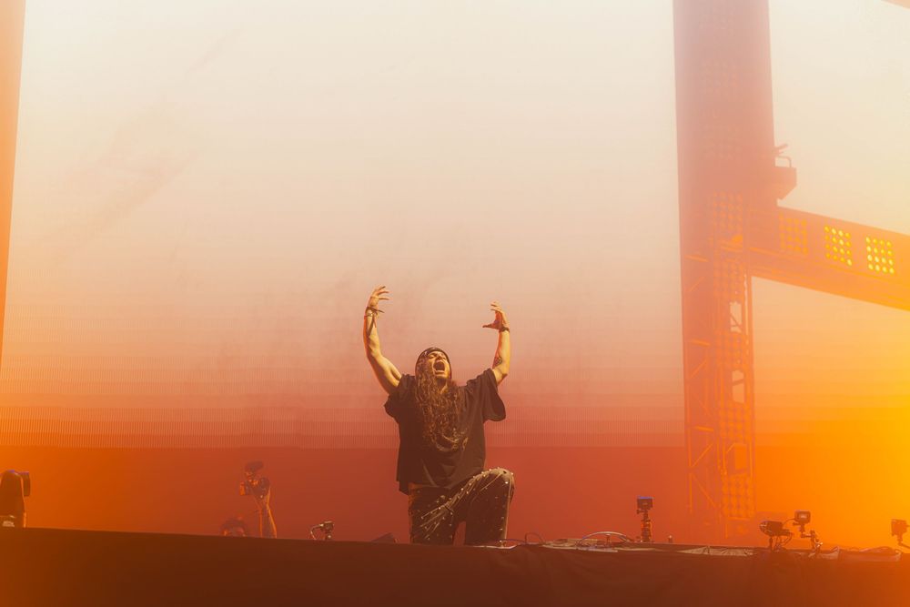 Concert crowd silhouettes raise their arms against a vibrant orange sunset sky during an outdoor music festival.