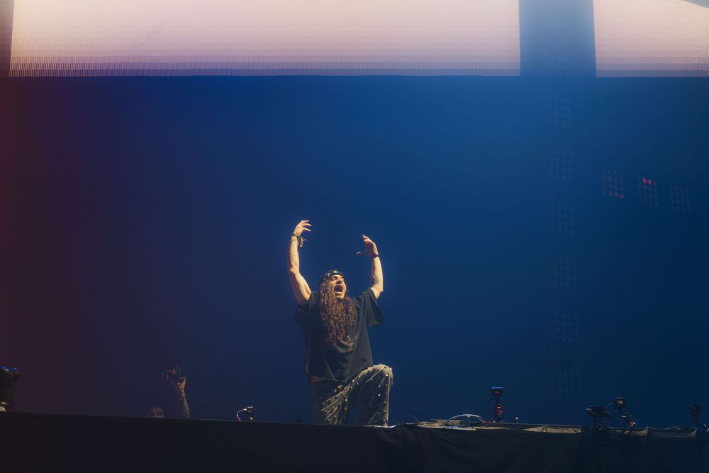 DJ performs on stage with raised arms while colorful lights illuminate the dark concert venue.