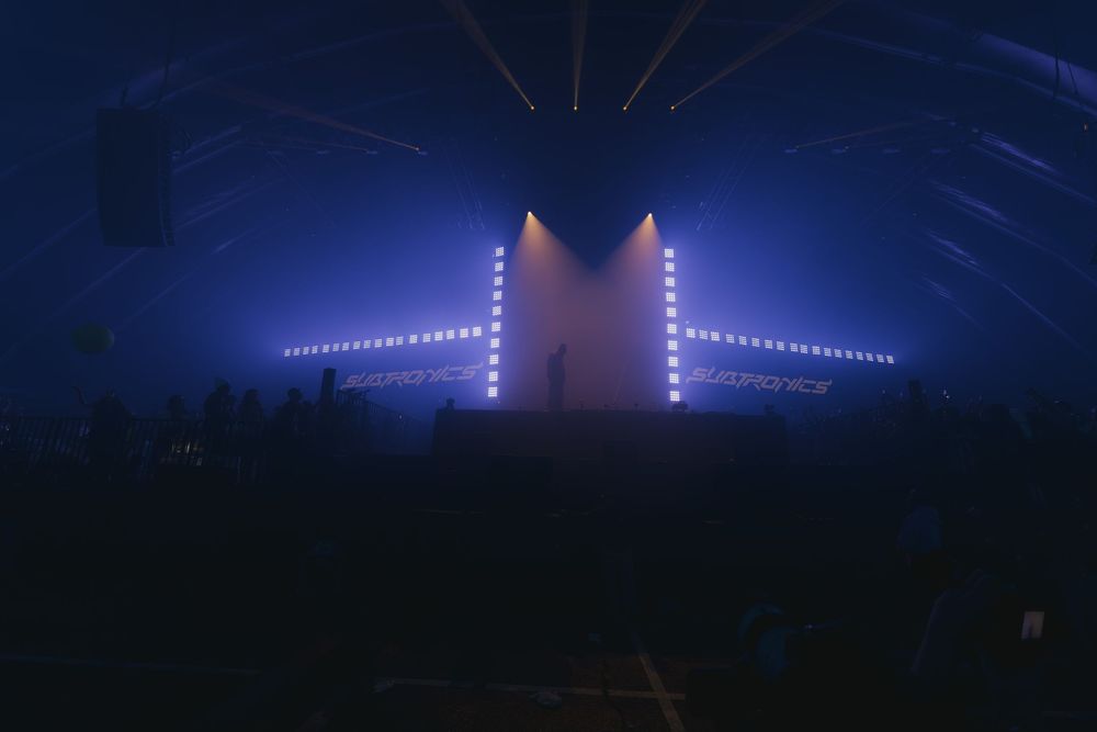 Concert stage illuminated with blue lights and vertical light beams creating a dramatic nighttime atmosphere.