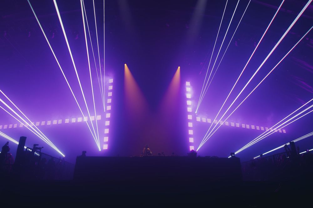 Dramatic purple concert stage lighting with vertical laser beams and fog creating an atmospheric nightclub environment.