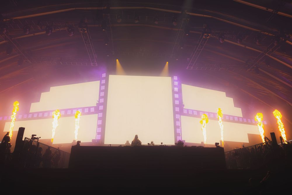 Music festival stage bathed in warm sunset colors with vertical light beams creating an atmospheric panoramic view.