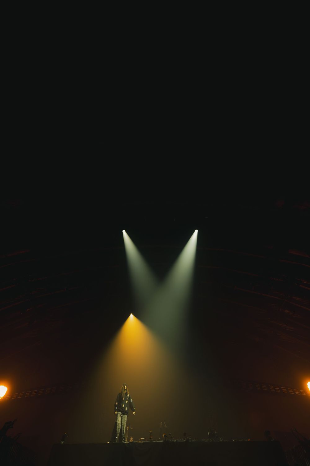 Silhouetted performer stands on dark stage illuminated by dramatic yellow and green spotlights.
