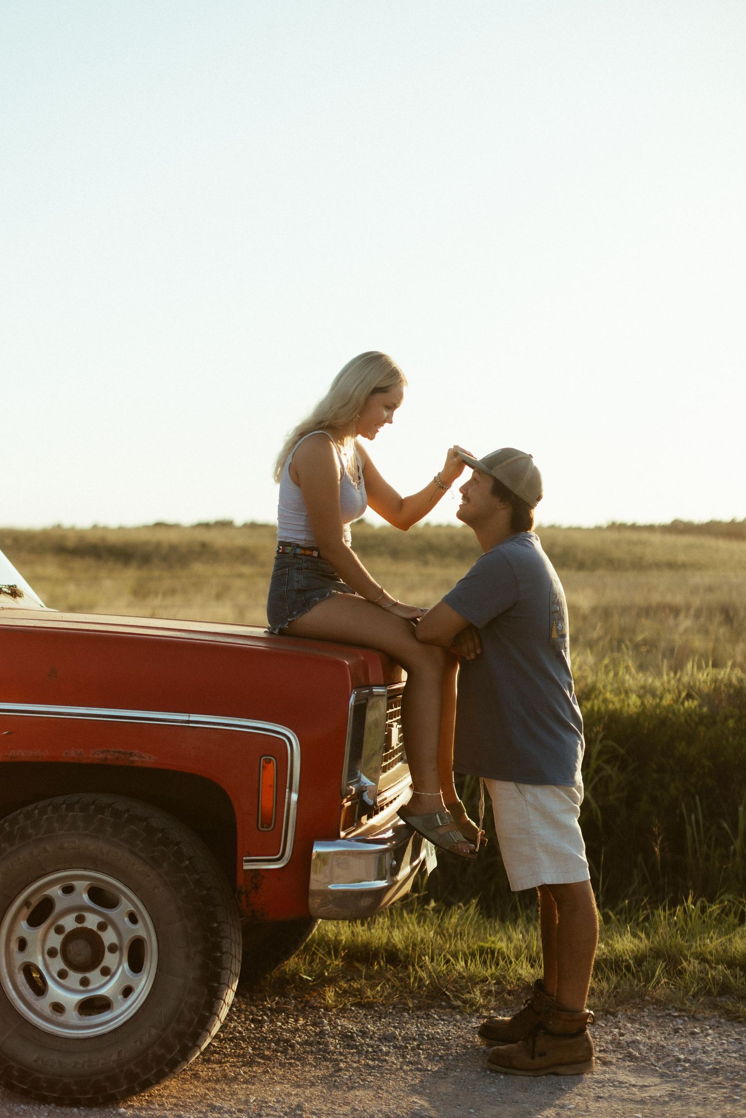 A couple shares a romantic moment at sunset next to a vintage red pickup truck on a dirt road.