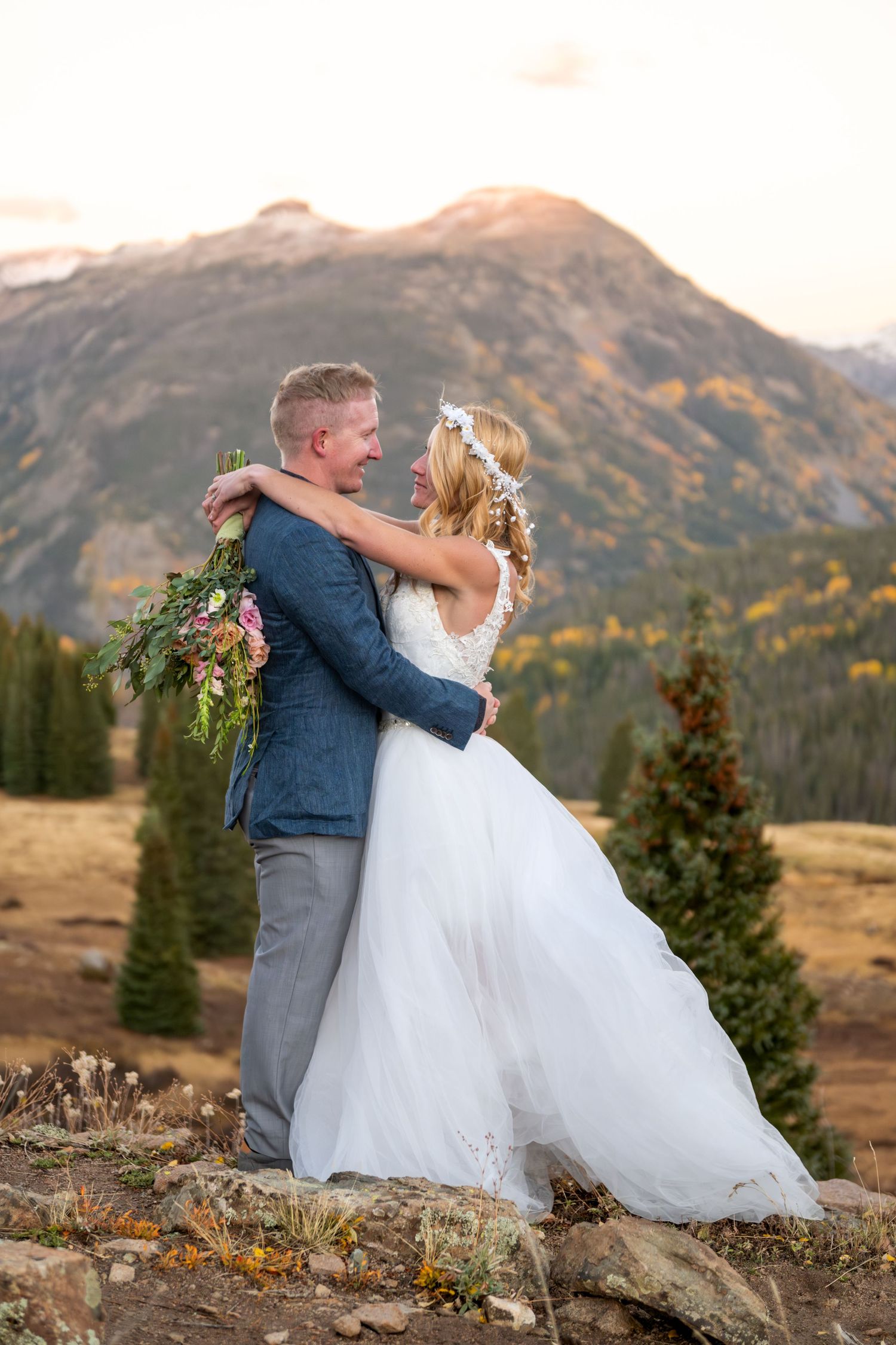 A bride and groom share a hug with the dramatic peaks of the San Juan mountains in alpenglow behind them. She is in a flowy white dress.