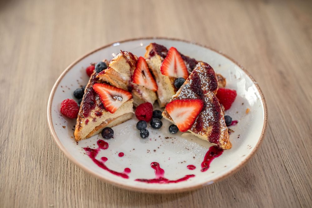 French toast topped with fresh strawberries, blueberries, and bacon on a white plate with raspberry sauce drizzle.