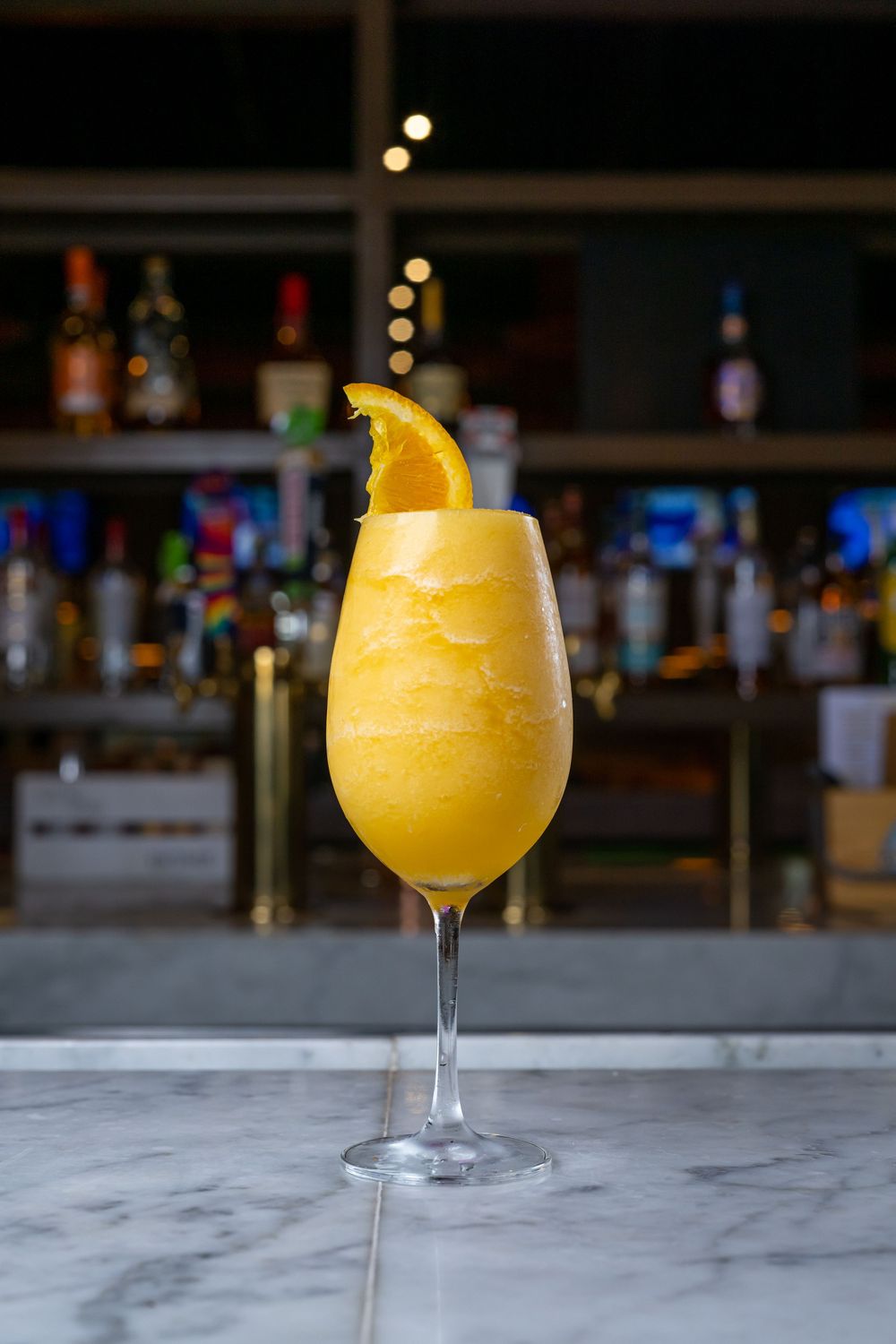 Four identical yellow frozen cocktails in wine glasses sitting on a marble bar counter with blurred background lighting.