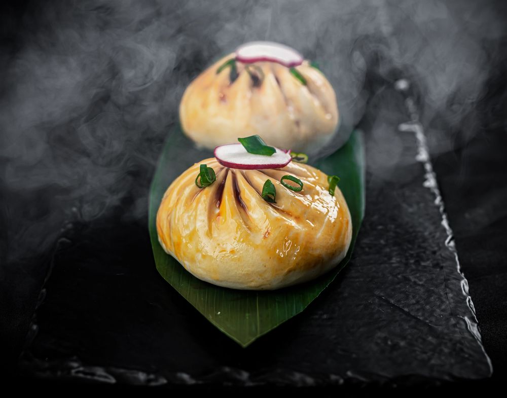 Two steamed Asian dumplings served on green leaf against dark stone surface with dramatic steam effect.