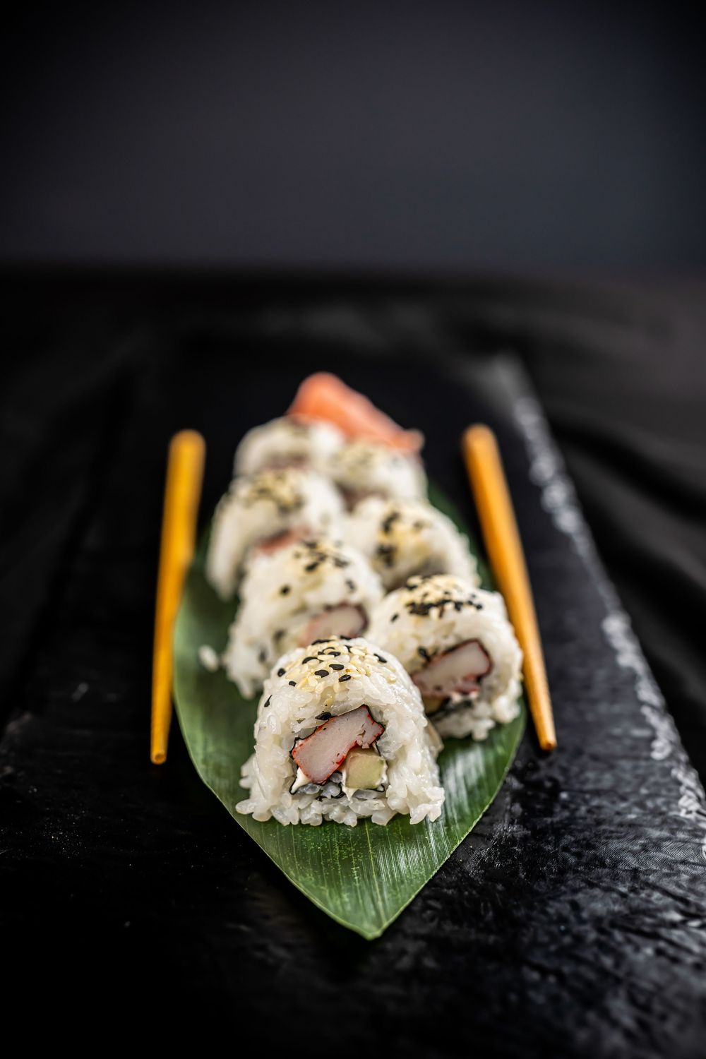 Close-up sequence of California sushi roll with black sesame seeds on dark slate platter with bamboo chopsticks.