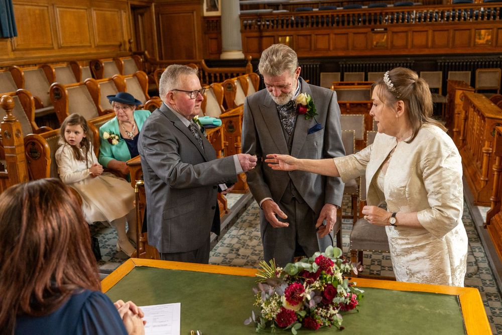 The best man hands the grooms wedding ring to the bride during their Southport Town Hall ceremony, with family watching from the seats.