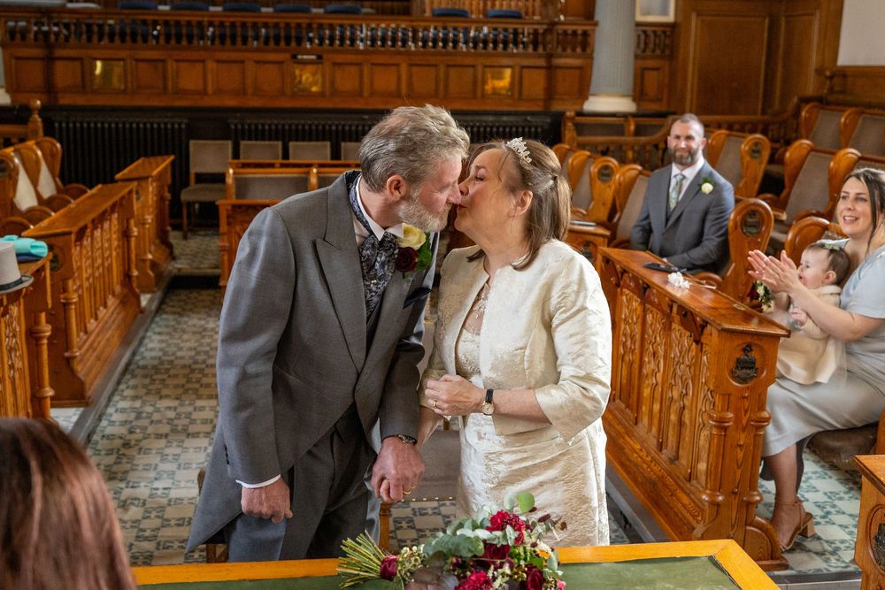 Bride and groom share their first kiss after the ceremony at Southport Town Hall, with family clapping in the background.
