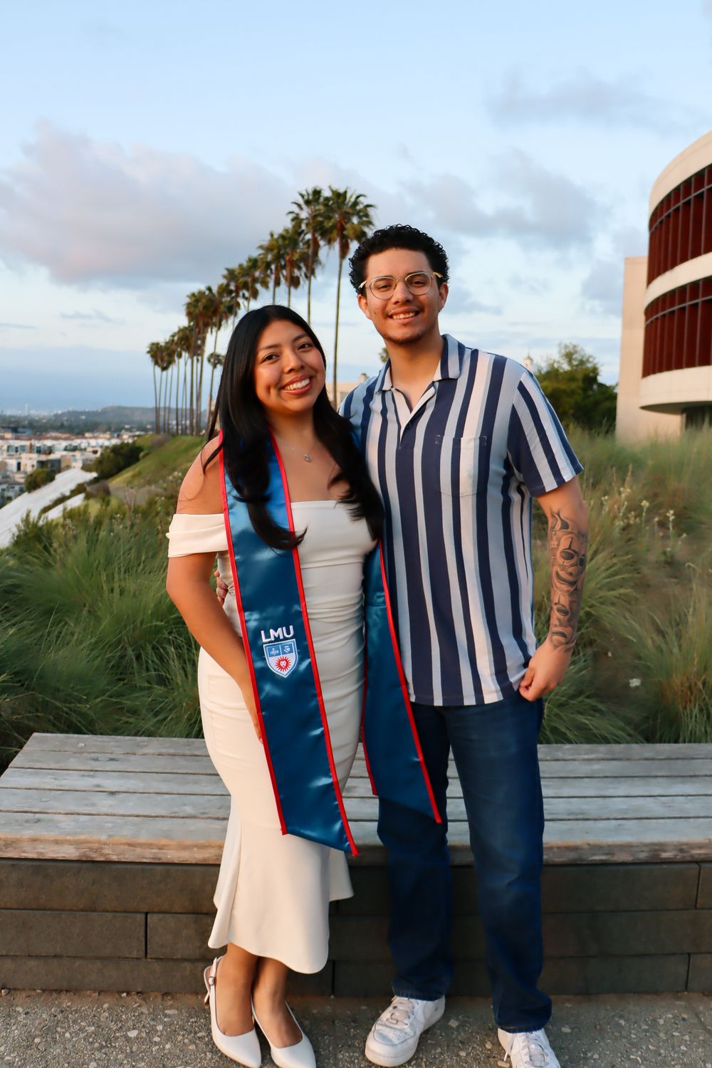 A couple poses together on a beachside walkway with palm trees and ocean views at sunset.