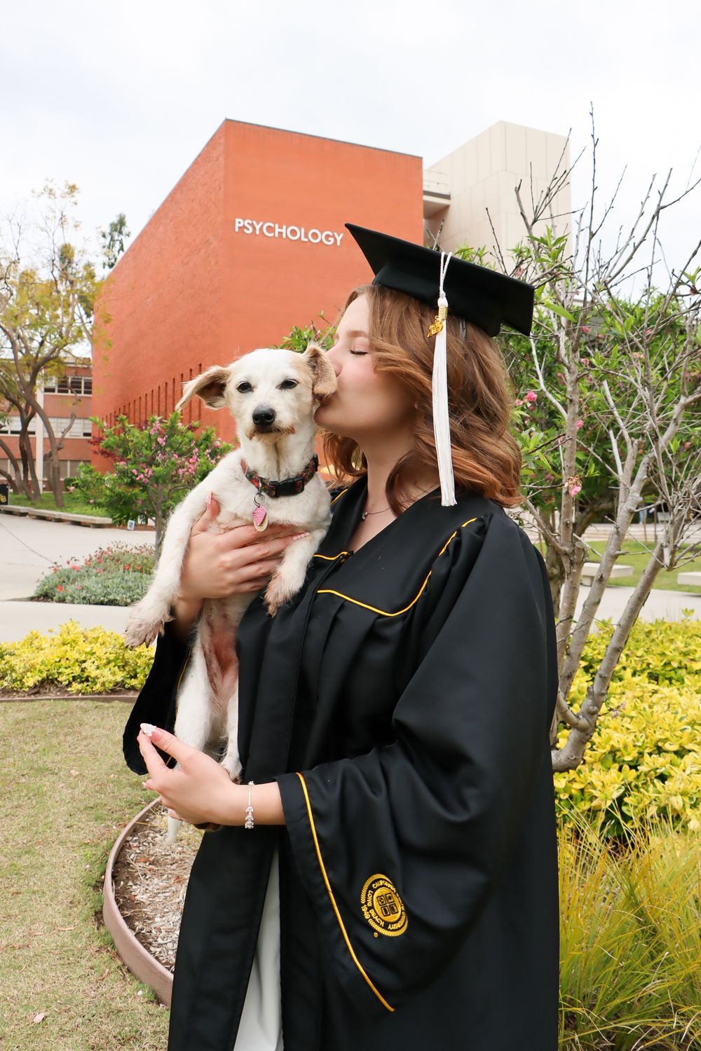 Graduate in black cap and gown holds and kisses small white dog during outdoor graduation photo session.