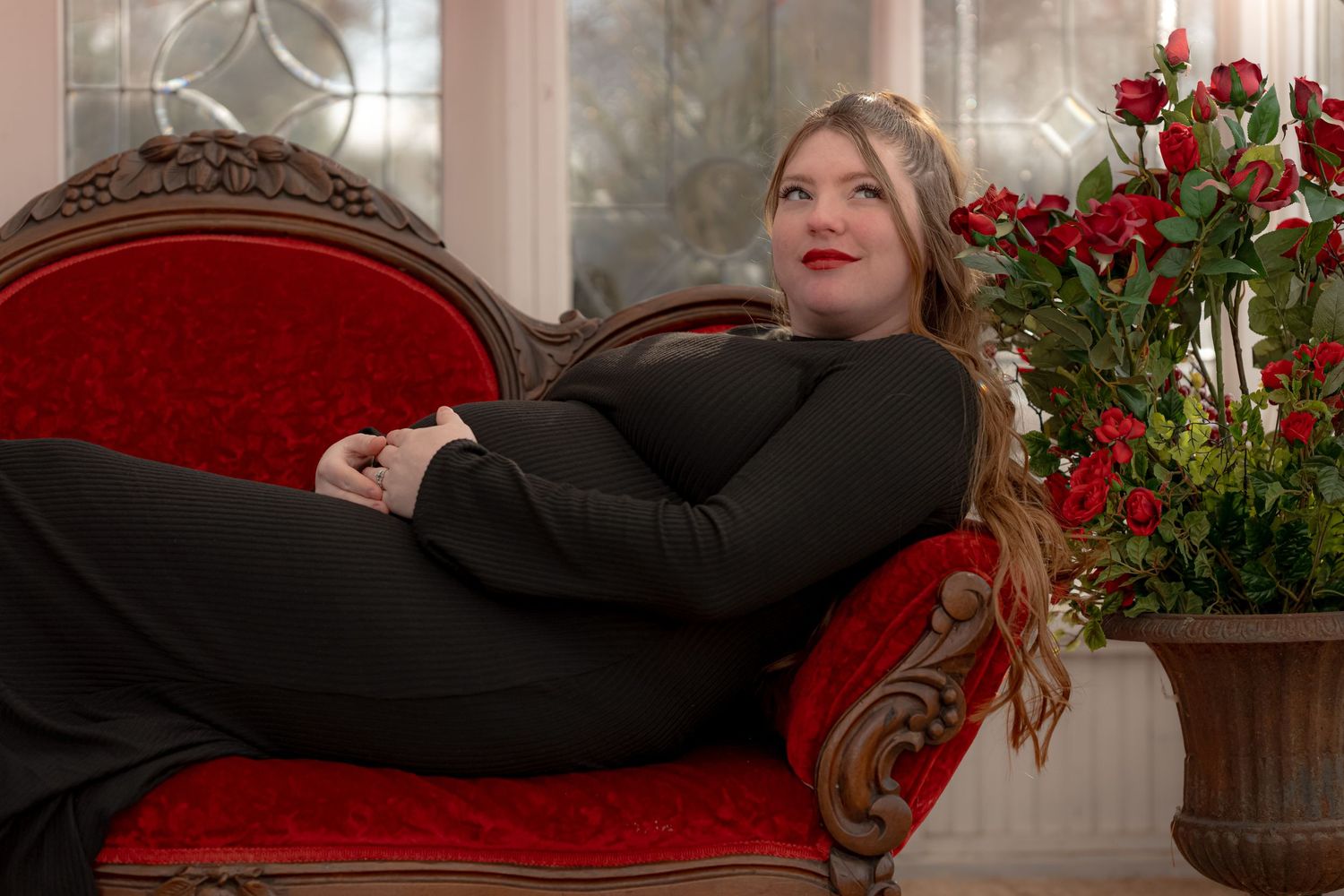 Woman in black dress lounges on red velvet chaise lounge with red roses in elegant interior setting.