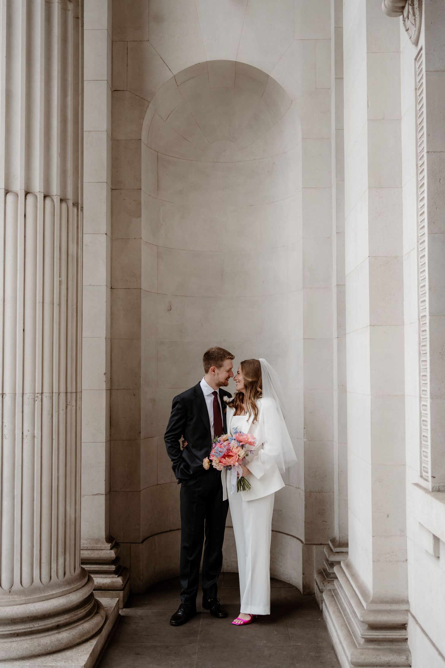 Elegant wedding couple embraces between white marble columns in a classical architectural setting.