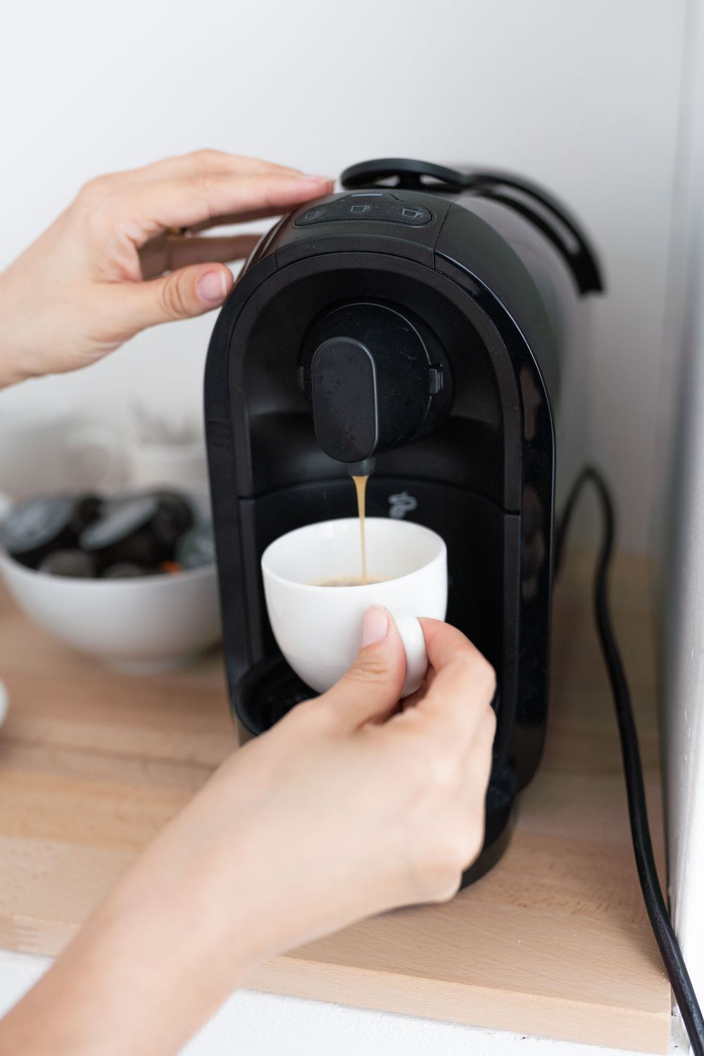 A sequence of hands operating a black coffee maker machine to brew and dispense coffee into a white cup.