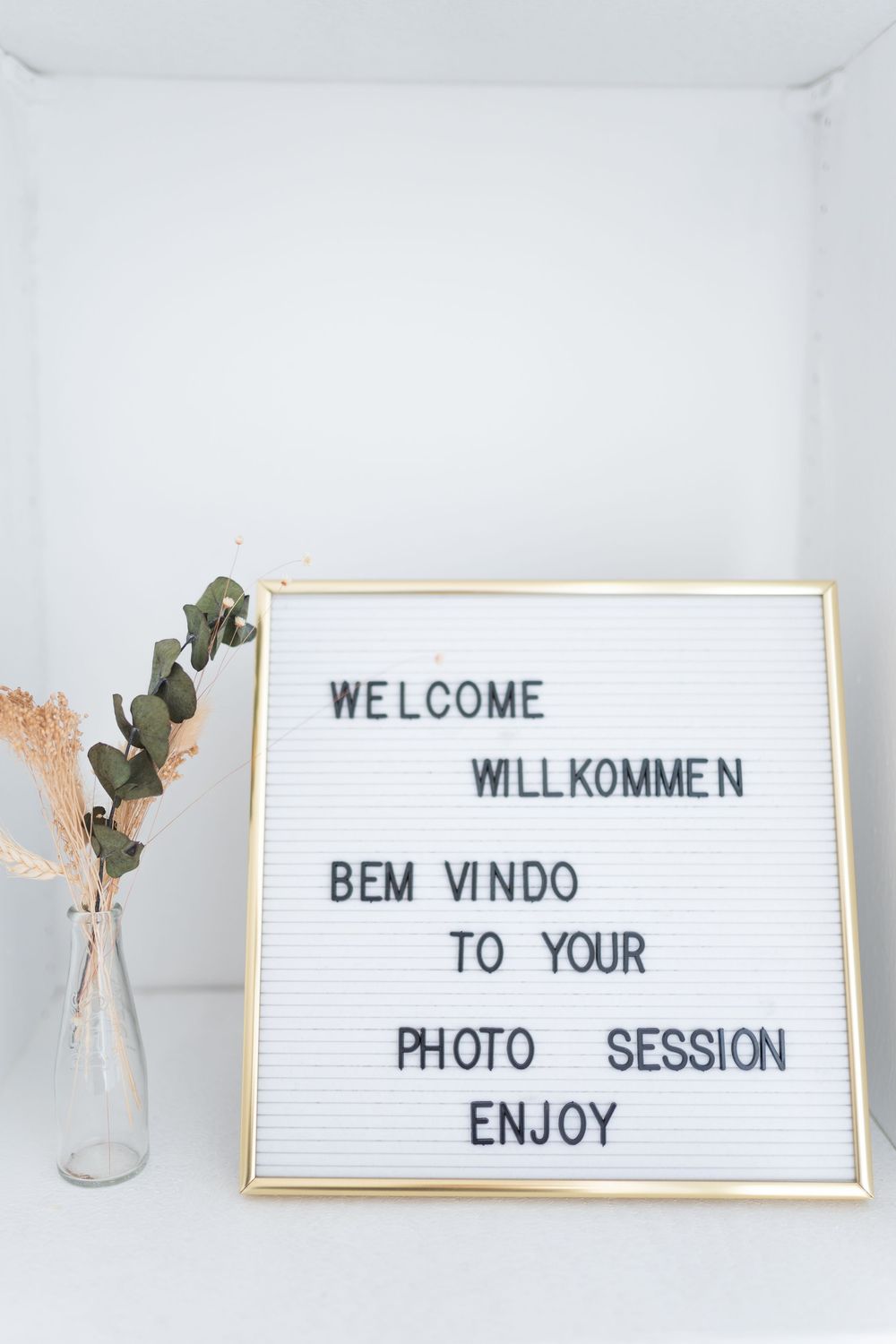 Letter board welcomes guests to photo session in multiple languages with eucalyptus branch decor on white surface.