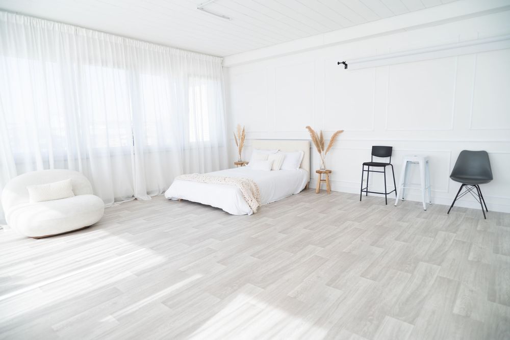 Modern minimalist bedroom interior with white walls, light gray wood flooring, and soft natural lighting.