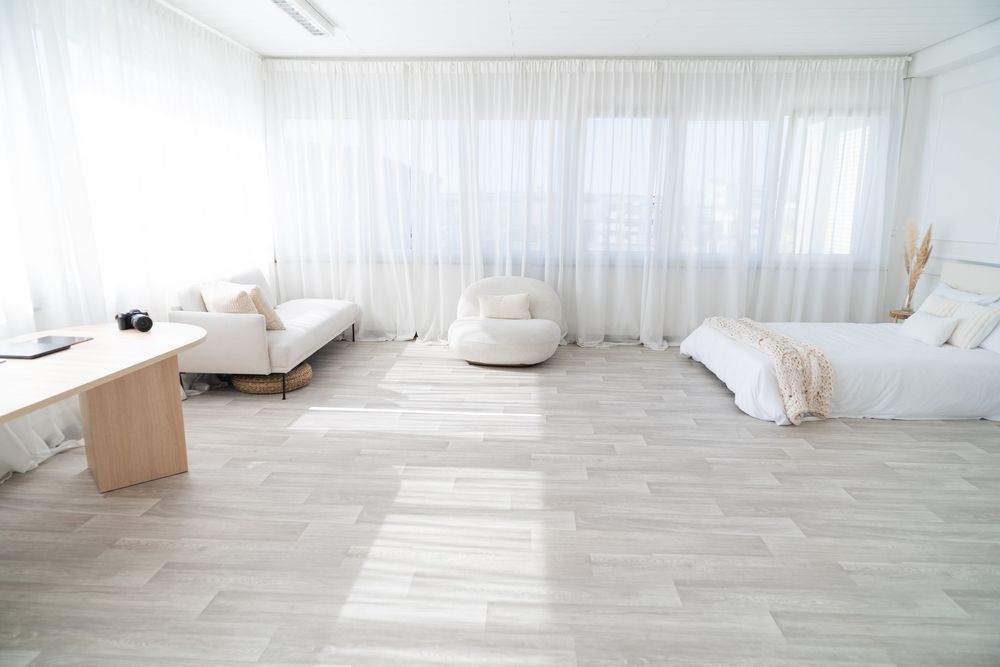 Minimalist white studio apartment interior with light wood floors, sheer curtains, and modern furnishings.