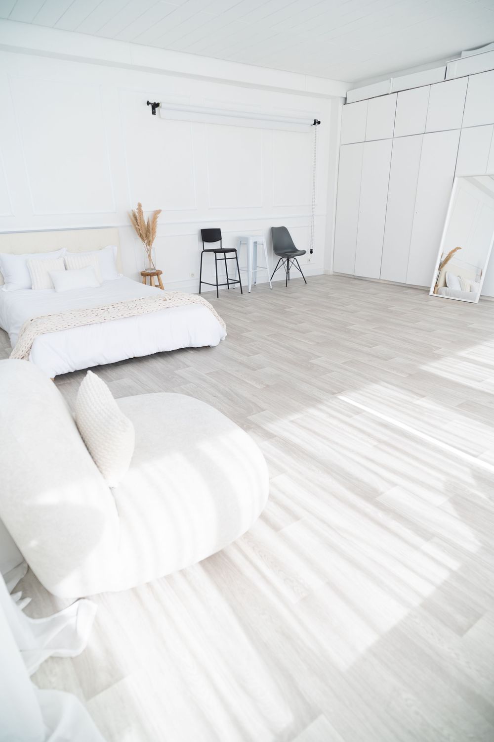 Minimalist white bedroom studio space with light wood floors, white bedding, and modern accent chairs.