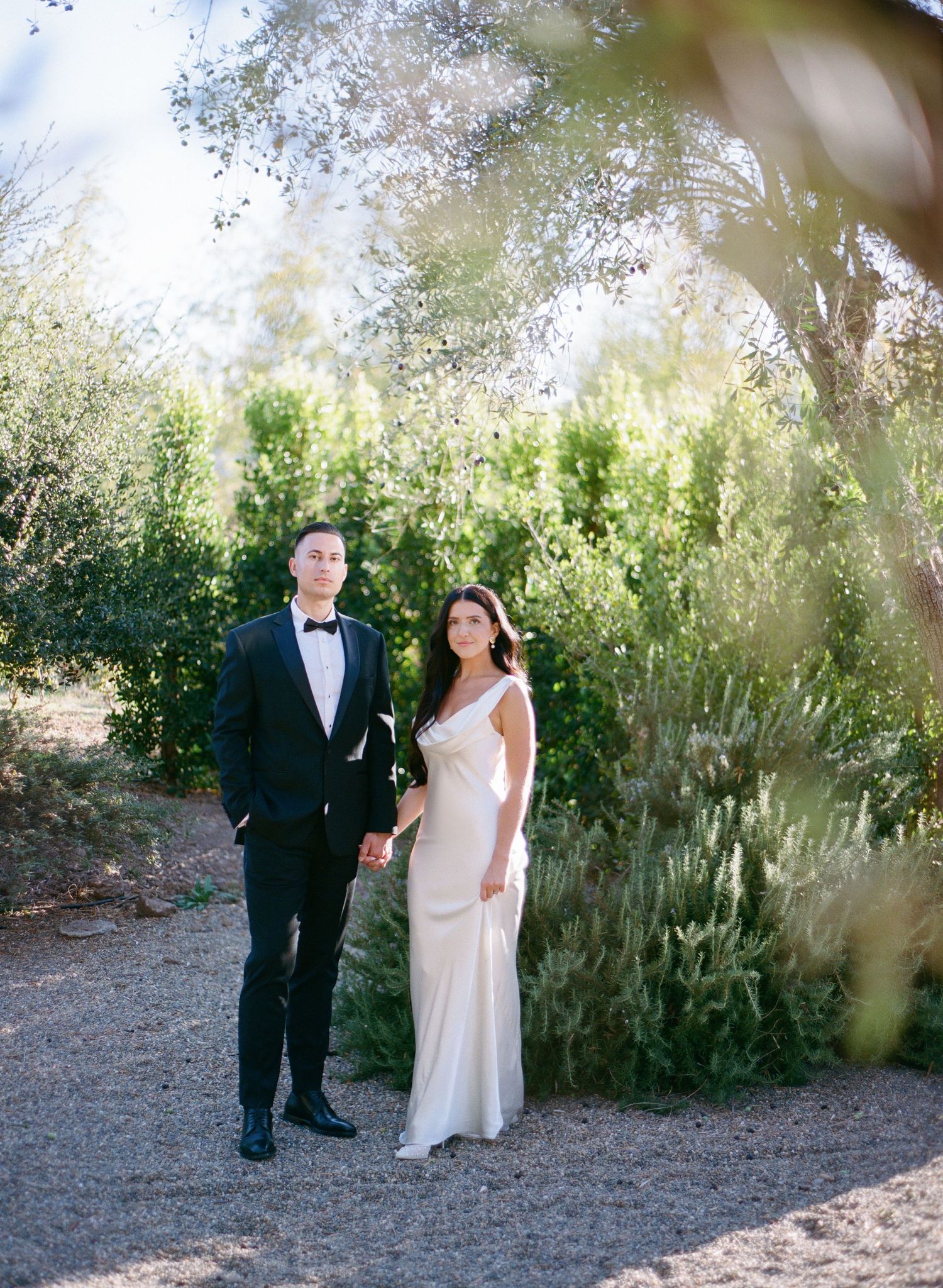 A couple walks together in a garden setting with natural foliage and sunlight filtering through trees.