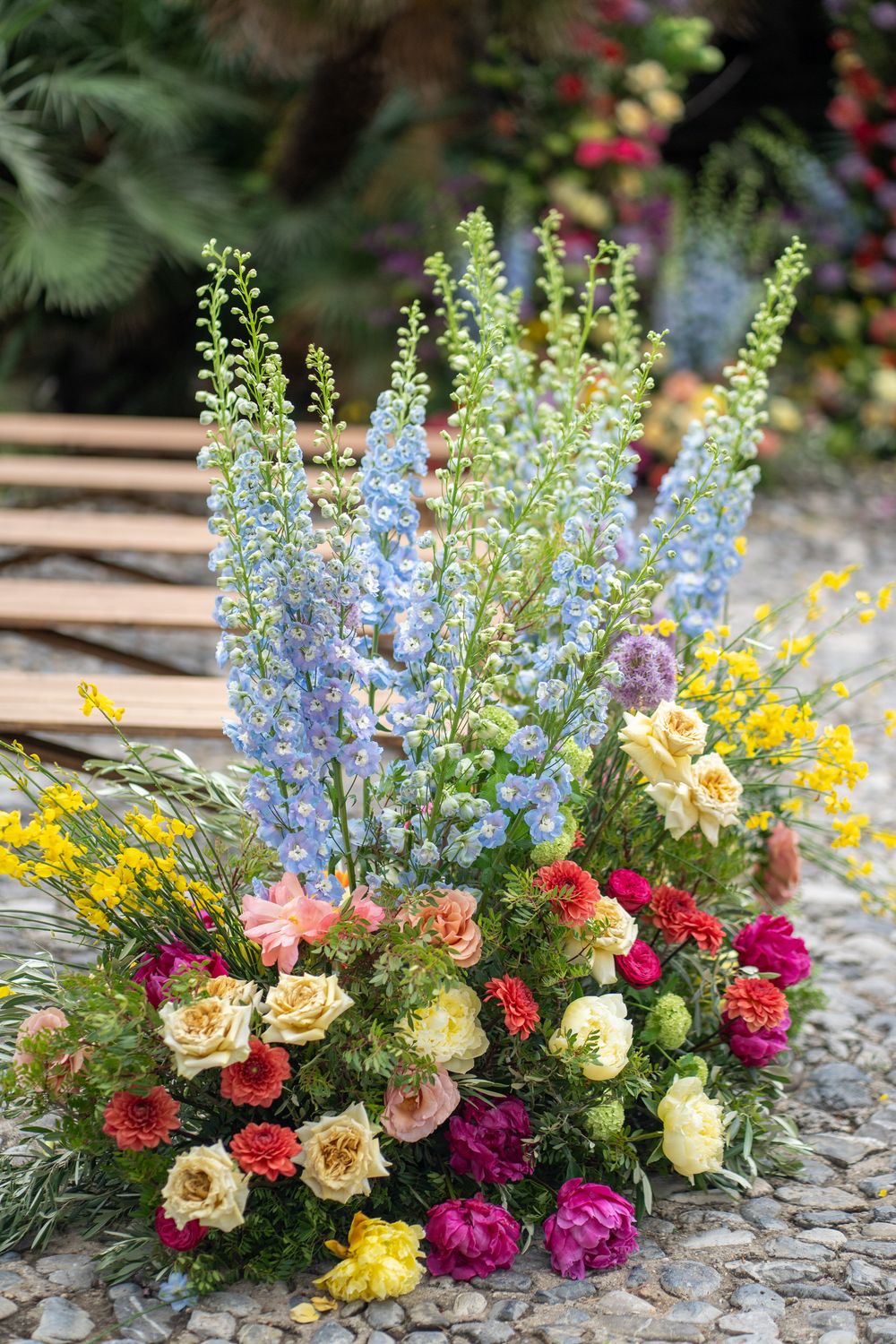 A colorful floral arrangement with roses, delphiniums, and mixed blooms displayed on a stone surface in natural light.