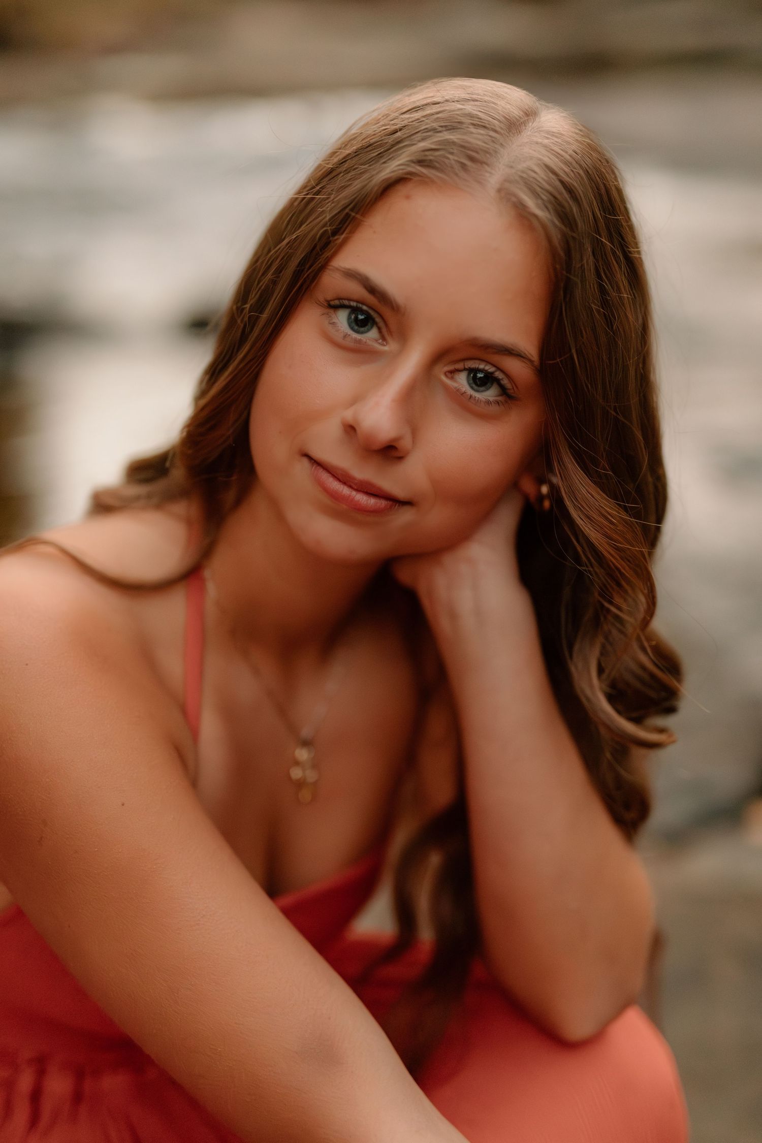 Beautiful young woman with green eyes and wavy brown hair posing in a coral dress near water.