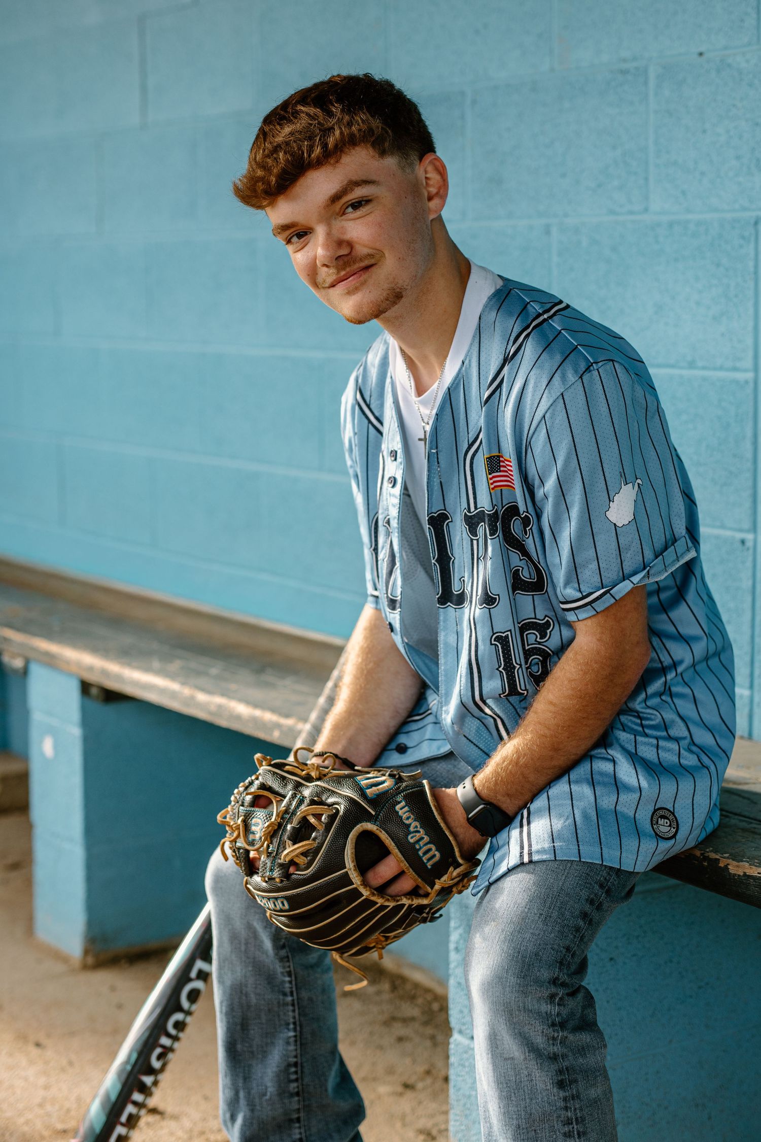Young baseball player in blue pinstripe jersey holding glove sits in dugout smiling.