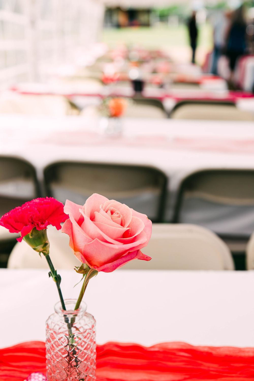 A pink rose and red carnation in a crystal vase on a white tablecloth at an event.