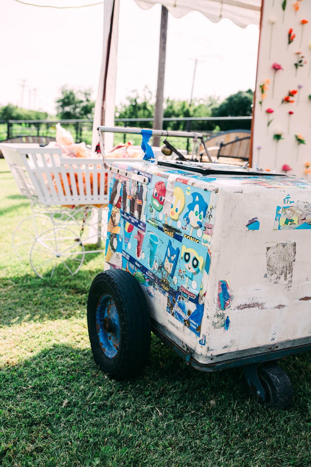 A vintage ice cream cart decorated with colorful stickers sits on grass at an outdoor event.