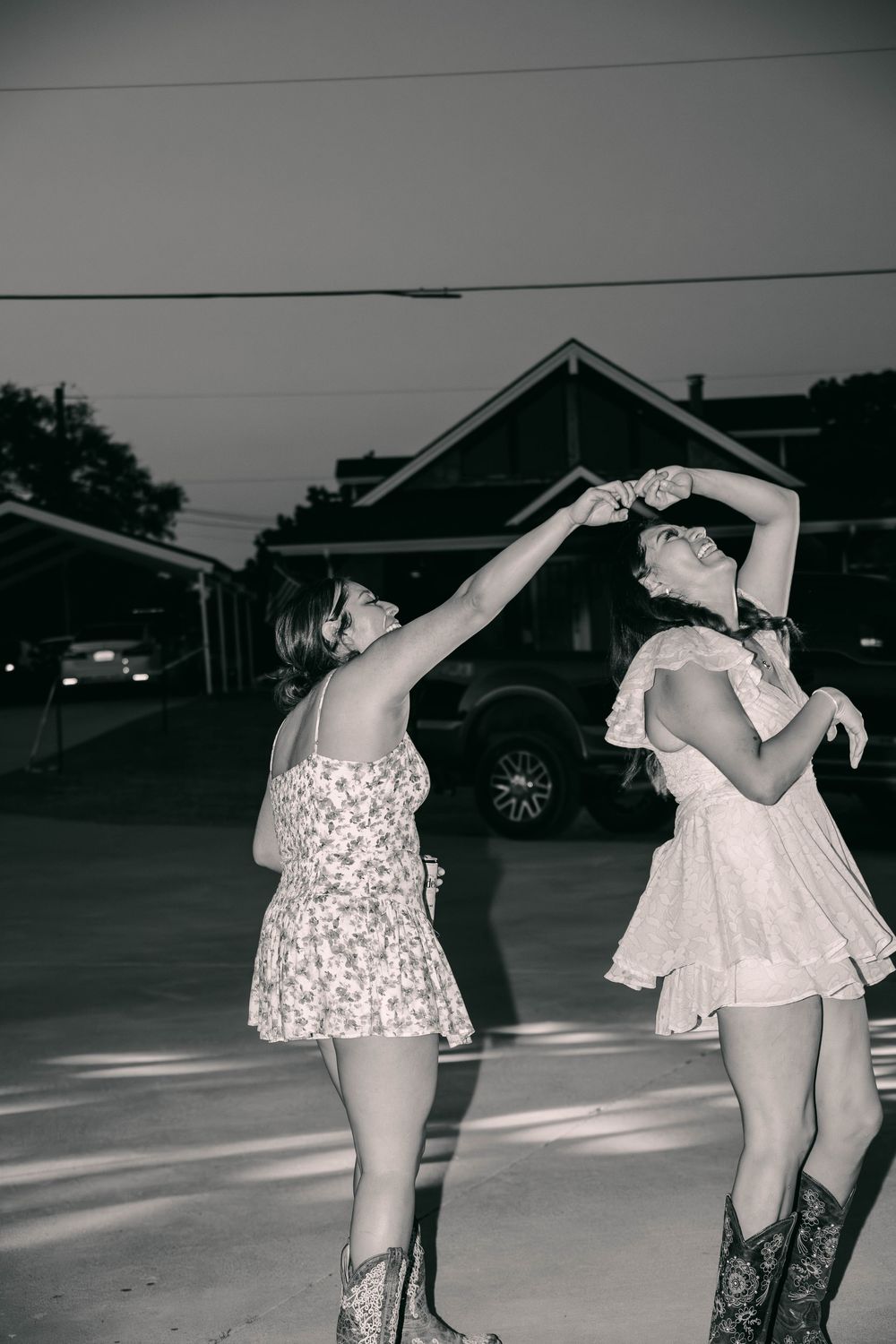 Two people in floral dresses dancing together in a parking lot at dusk.